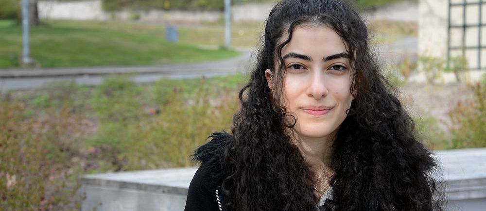 Person med långt lockigt hår sitter utomhus på en bänk, grönska i bakgrunden.