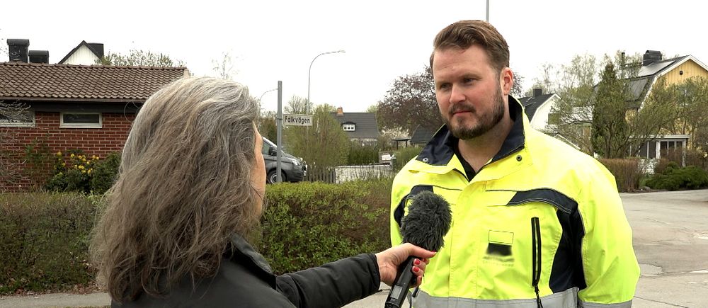 En kvinna med gråsprängt hår intervjuar en man med kort brunt hår ikväll varseljacka. I bakgrunden ett bostadsområde med villor.