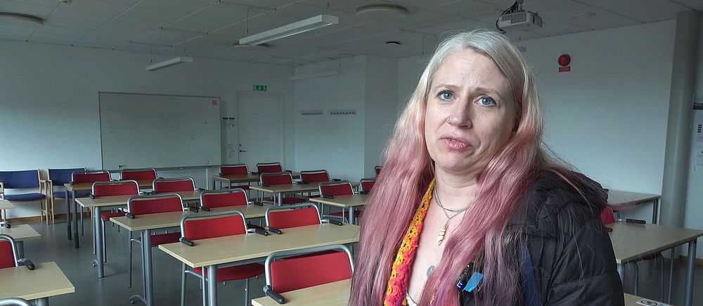 Kvinna med långt hår står i ett tomt klassrum med rader av skrivbord och stolar.
