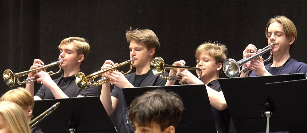 FEM UNGDOMAR STÅR OCH SPELAR TRUMPET I ETT STORBAND.