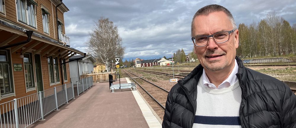Man i randig tröja står vid en tågstation med spår och byggnad i bakgrunden.