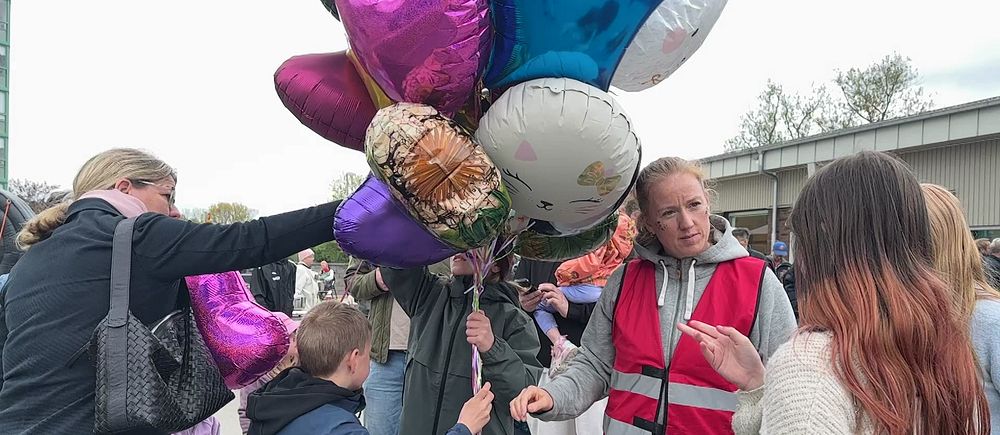 Person i röd väst delar ut heliumballonger till barn och vuxna vid en utomhusaktivitet.