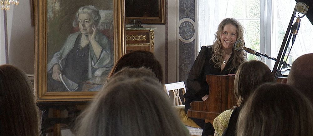 Sångerskan Sofia Karlsson spelar piano inför publik, ett stort porträtt av Selma Lagerlöf syns bredvid henne.