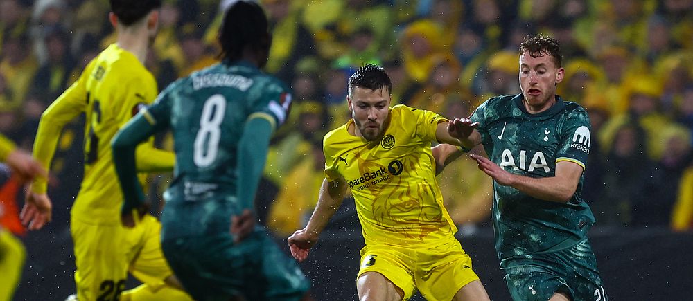 Tre fotbollsspelare kämpar om bollen under en match mellan lag i gult och grönt.
