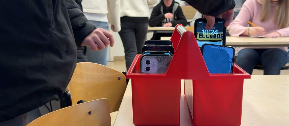 Mobiltelefoner samlade i en röd låda i ett klassrum. Elever i bakgrunden.