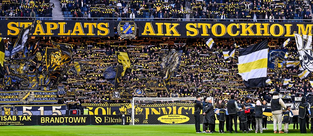 Fullsatta AIK-läktare med svartgula flaggor och banderoll med texten ”Ståplats AIK Stockholm”.