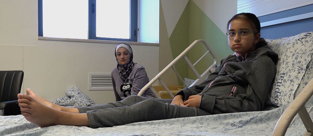 Amira ligger i sjukhussäng, hennes mamma sitter bredvid sängen.