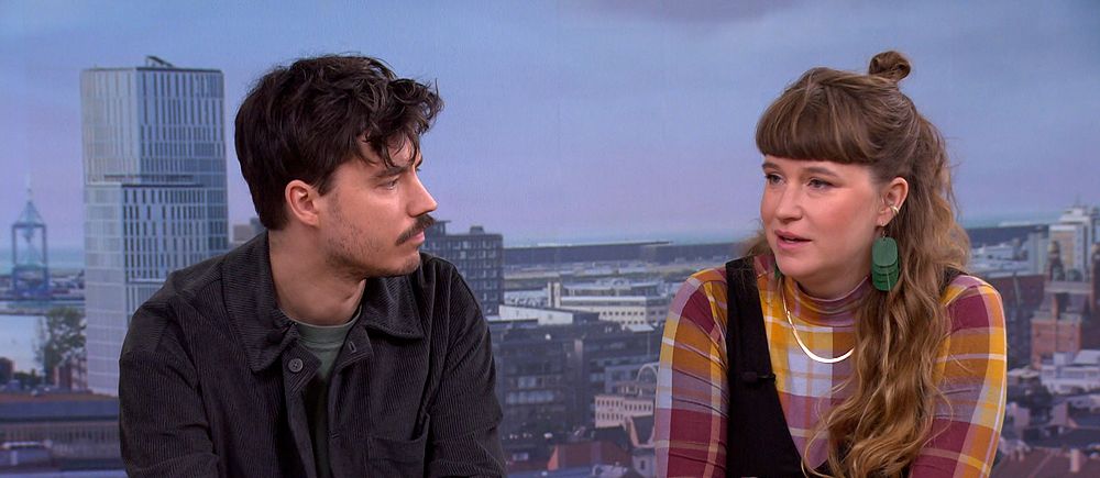 Två personer sitter vid ett bord i en tv-studio med stadsvy i bakgrunden.