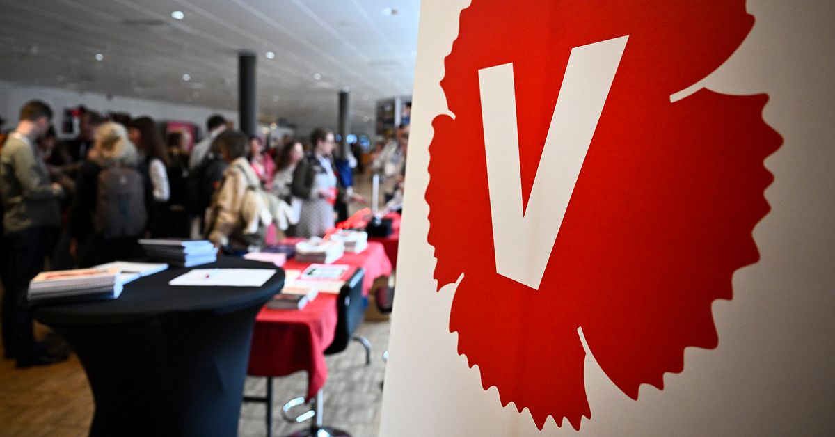 Vänsterpartiets röda V-logga på vägg i möteslokal med folk i bakgrunden