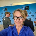 En person tar en selfie med NATO-logotyper i bakgrunden. Fyra personer syns stående, vända bort från kameran, i en professionell miljö. Det verkar vara en presskonferens eller ett evenemang kopplat till NATO.