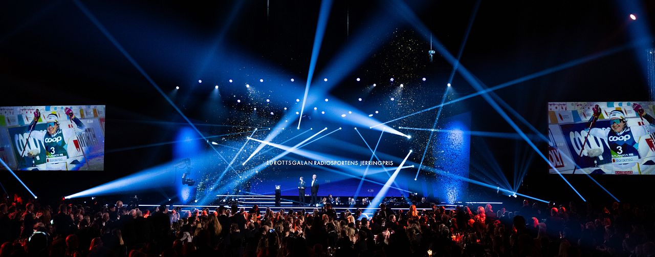 En scen i en stor evenemangsarena med starka ljusstrålar som strålar från taket. Publiken är samlad och delvis i mörker, med skärmar som visar bilder på scenen. Eventet verkar vara en prisutdelning eller gala, med diplomör och festlig atmosfär.