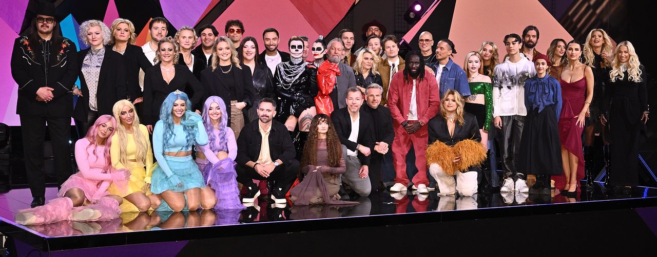 Gruppbild av deltagare och artister från Melodifestivalen 2025, klädda i färgglada och varierande kläder, poserande på en scen med geometriska bakgrundselement.