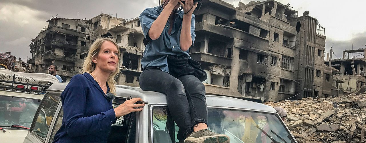 Stina Blomgren och fotograf Pernilla Edholm i en krigshärjad stad. Pernilla sitter på taket på en bil.