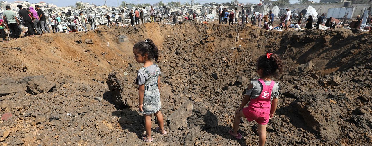 Två barn står i en stor bombkrater omgivna av jord och ruiner, människor i bakgrunden. Bilden är tagen i Deir el-Balah i Gaza.
