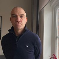 Stefan Dalteg står vid ett fönster med utsikt över stadsmiljö. Han tittar allvarligt in i kameran.