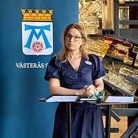 Två personer står vid talarstolar, Västerås stadsvapen syns i bakgrunden.