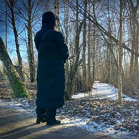 Person i lång kappa står vid snötäckt stig i skog med solljus i bakgrunden.