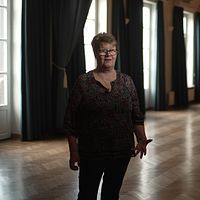 Finska föreningen i Stockholms ordförande Seija Sjöstedt intervjuas i mörkt rum med stora fönster och långsträcka gardiner.