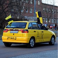 Delad bild. En gul bil med gulsvarta Skellefteå AIK-flaggor och text på. Moderata politikern Andreas Löwenhöök iklädd svartgul slips och kavaj.