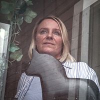 Karolin Norlén Järpeskog tittar ut genom fönstret på ett HVB-hem. Inomhusmiljö med gröna växter, en dalahäst och träpanel i bakgrunden.
