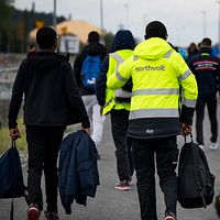 Flera Northvoltarbetare med väskor promenerar längs en väg, några bär reflexjackor där det står Northvolt på ryggen.