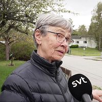 Reporter håller SVT-mikrofon mot en äldre kvinna, polis står bakom avspärrning på lugn villagata.