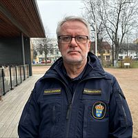 Polis i uniform står framför en parkmiljö med bostadshus i bakgrunden.