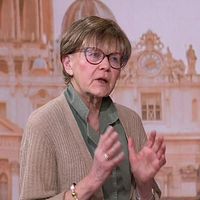 Person gestikulerar framför en bakgrund med klassisk arkitektur och kupolprydda byggnader. I bild syns Vatikankännaren Ulla Gudmundsson.