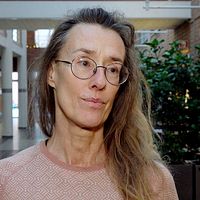 En person står inomhus med en växtfylld bakgrund av tegelväggar och en korridor. Personen bär en ljusrosa tröja med geometriskt mönster.