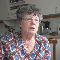 En äldre kvinna sitter i ett hemtrevligt kök med mikro och växter i bakgrunden.
