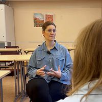 Två personer sitter vid skolbänkar och har ett samtal i ett klassrum.