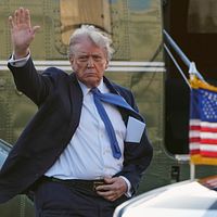 En man i kostym vinkar bredvid ett amerikanskt flagg framför ett helikopterplan.