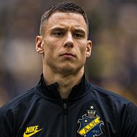 AIK-spelare med mörkt jacka och klubbmärke synligt, suddig publik i bakgrunden.