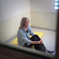 Kvinna sitter ensam på madrass i cell med kala väggar och fönster med galler.
