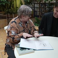 Två personer vid ett bord, diskuterar en bok med illustrationer.