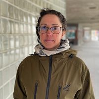 Person i grön jacka står i glasväggskorridor på trottoaren.