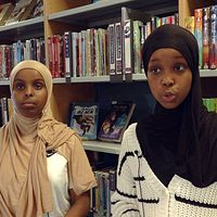 Två tjejer med hijab framför hylla med böcker i bibliotek