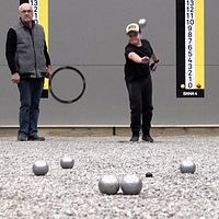 Två personer spelar boule inomhus mot en grå vägg med reklam.