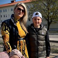Två personer står utomhus, en kvinna i gul klänning och ett barn i keps. Bakgrund med byggnad.