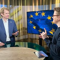 Alexandre Bloxs och Magnus Bergevin står vid ett bord i en studio. Tv-skärmen bakom visar EU-flaggan.