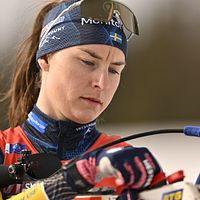 Skidskytt siktar med gevär under tävling, iklädd tävlingsdräkt och pannband.