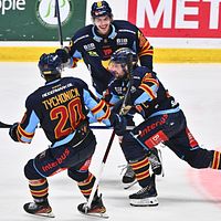 Tre hockeyspelare jublar och kramas på isen efter mål. Skridskor, klubbor och matchdräkter syns.