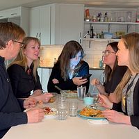 Fem personer samtalar runt ett bord i ett kök. En kvinna tittar på sin mobiltelefon.