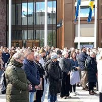 Stor folkmassa står samlad utanför en byggnad med svenska flaggor i solsken.