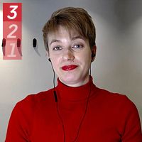 Person i röd tröja står i ett rum med enkel inredning och hylla på väggen.