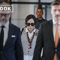 Flera personer i kostym och en kvinna med halsband och stora solglasögonen promenerar i tingsrätten.