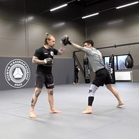 Två personer tränar kampsport i en gymmiljö. Flera andra syns i bakgrunden.