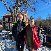 Man och kvinna står framför röd stuga i vintrigt landskap.