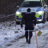 En person med en jacka där det står polis på ryggen, går mot en polisbil som står längre bort. På marken är det snö och runtomkring hänger avspärrningsband.