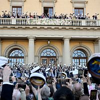 Studenter firar utanför skolbyggnad, viftar med mössor mot balkong.